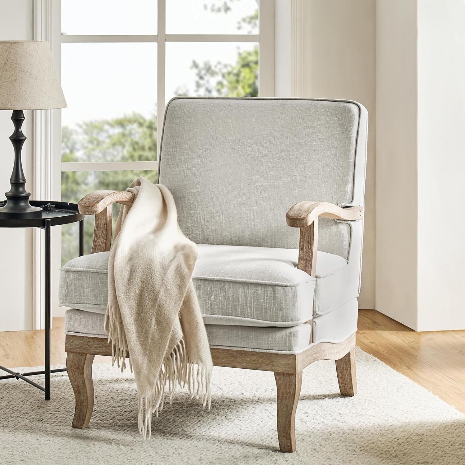 Gray upholstered armchair with wooden arms and legs, draped with a beige blanket, in a bright room.