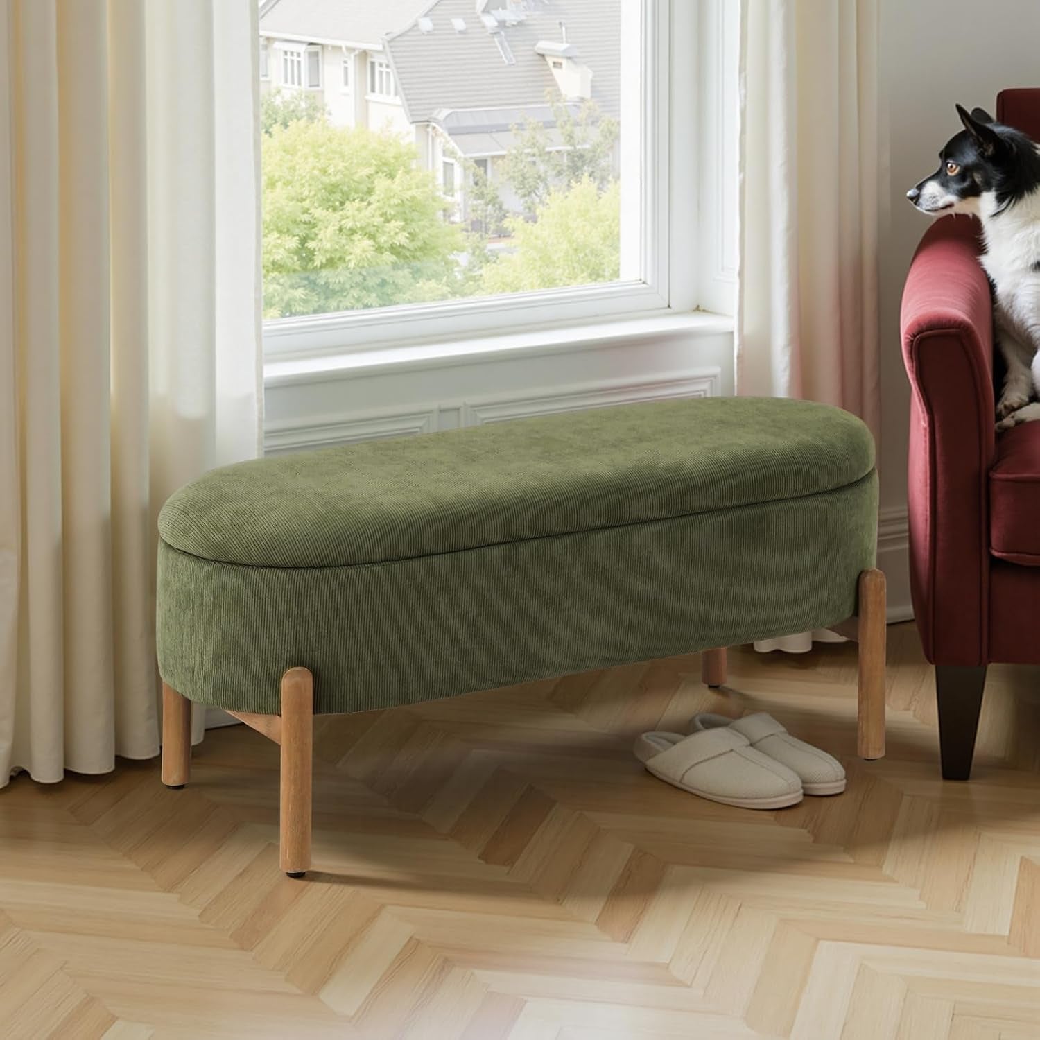 Green ottoman with wooden legs in a room with a window and a red chair.