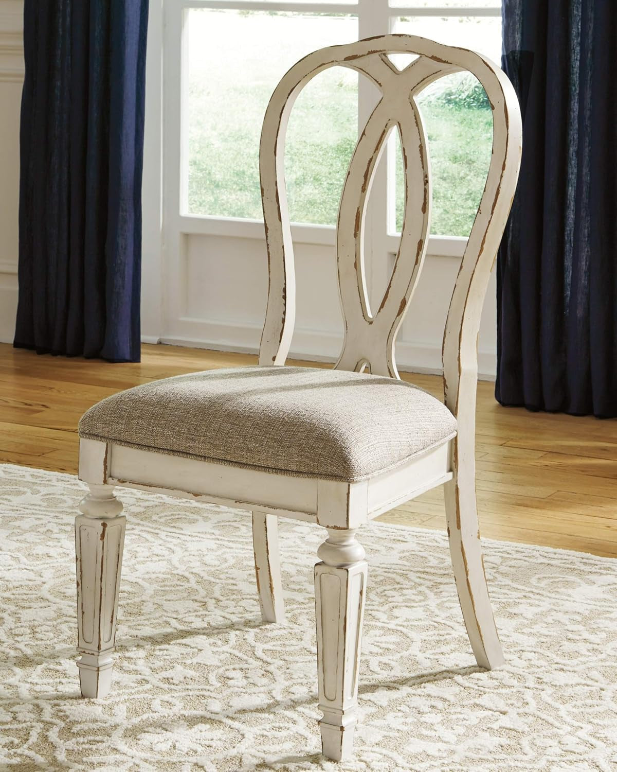 White wooden chair with a patterned cushion on a wooden floor with blue curtains in the background.