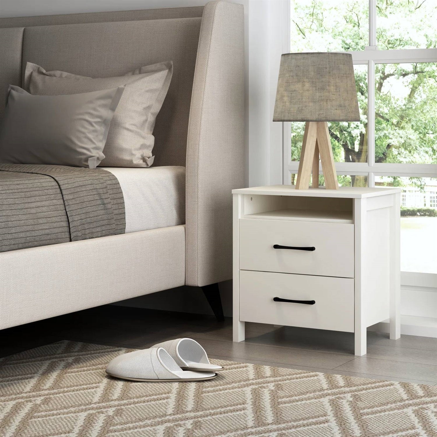 Neatly made bed with pillows, a nightstand with a lamp, and white slippers on a patterned rug.
