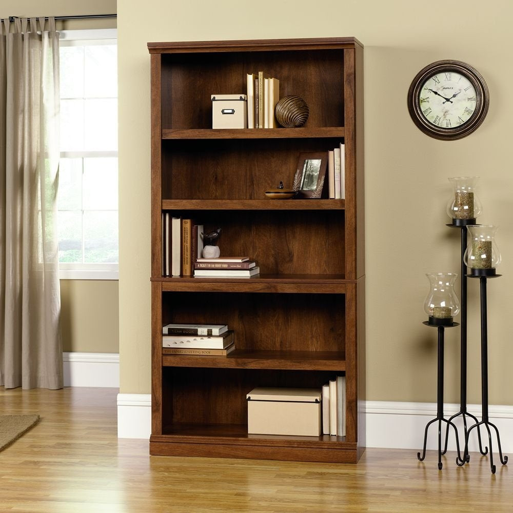 Wooden bookshelf with books and decor items in a room with a clock and lamp.