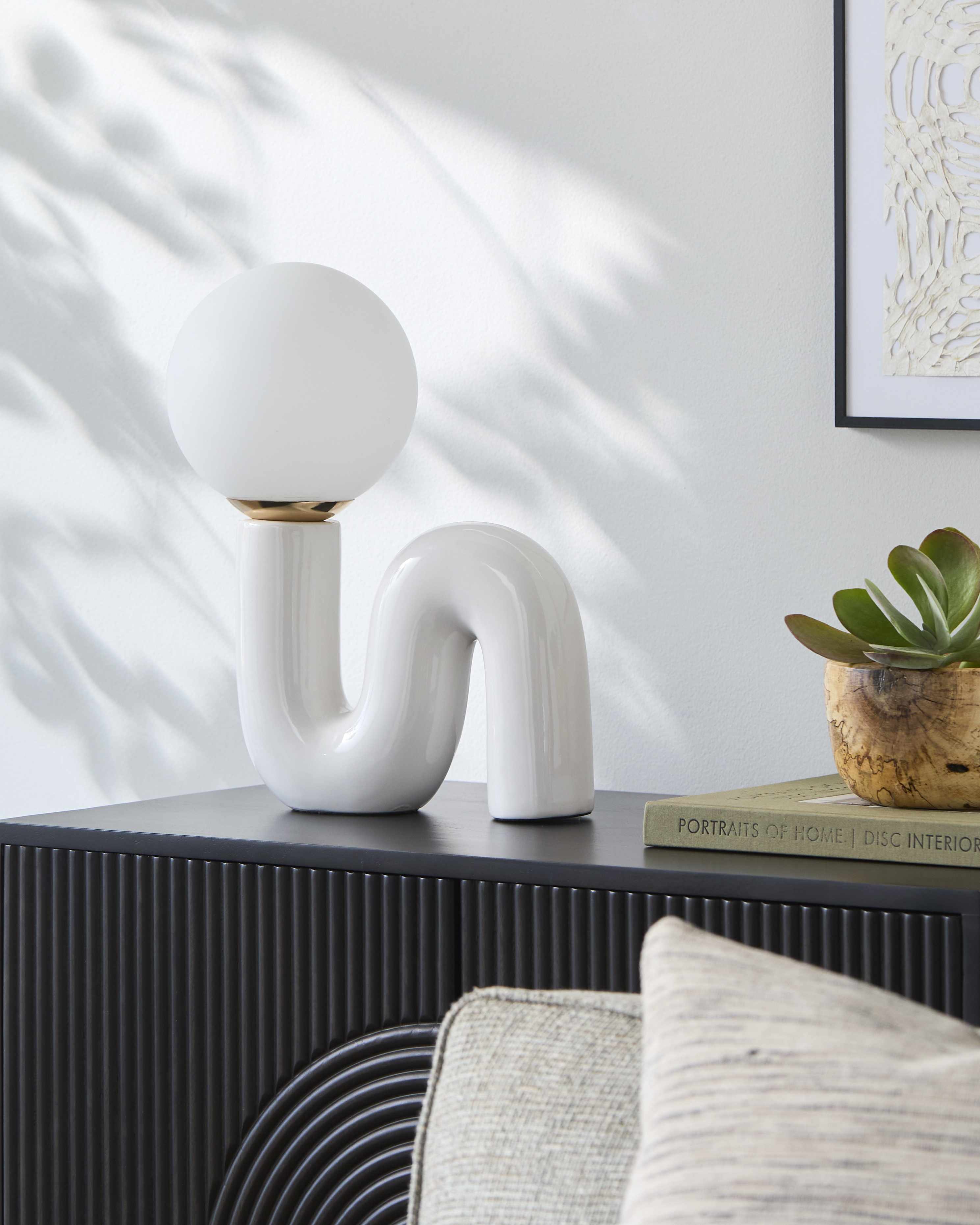 Modern white lamp on a dark surface with a light gray wall and framed artwork in the background.