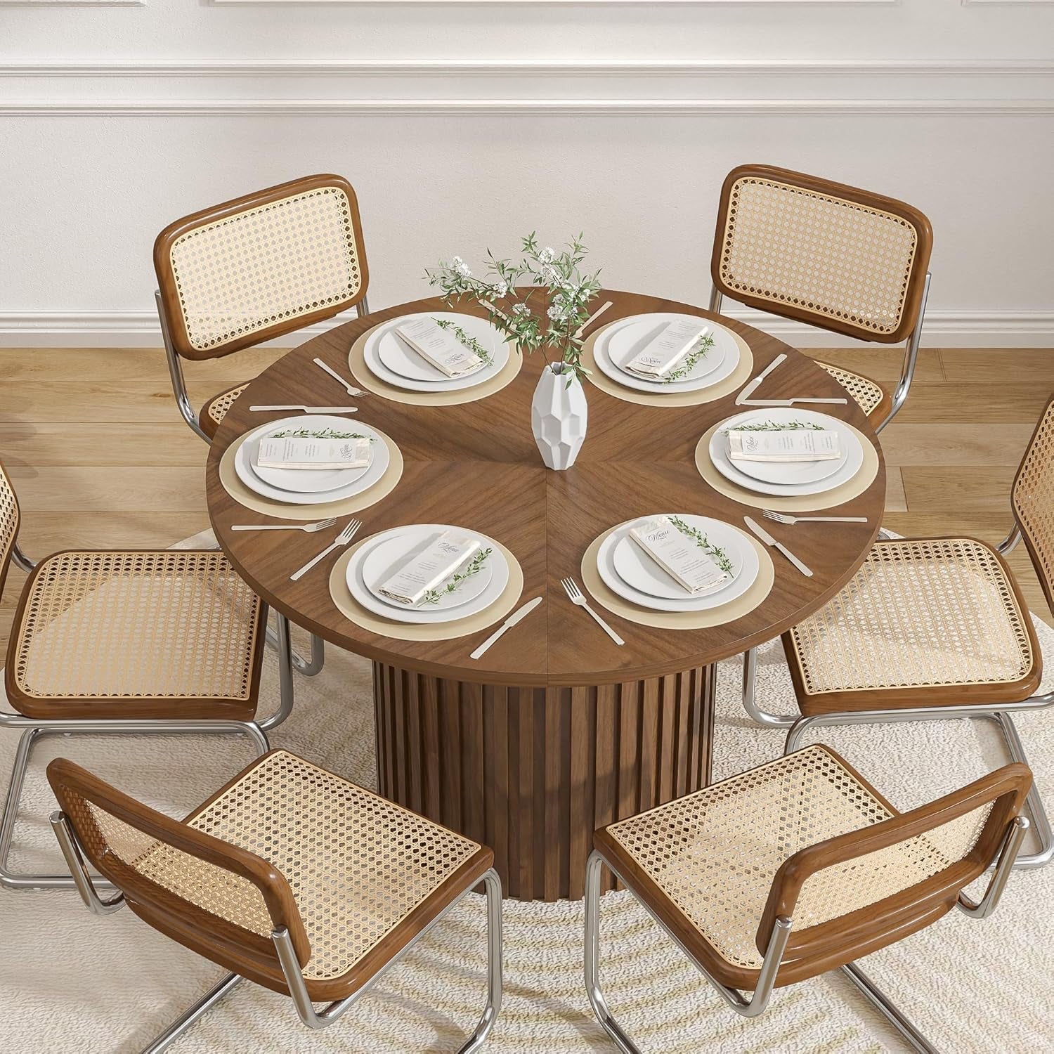 Dining table set with plates, cutlery, and a vase in a room with wooden flooring and white walls.