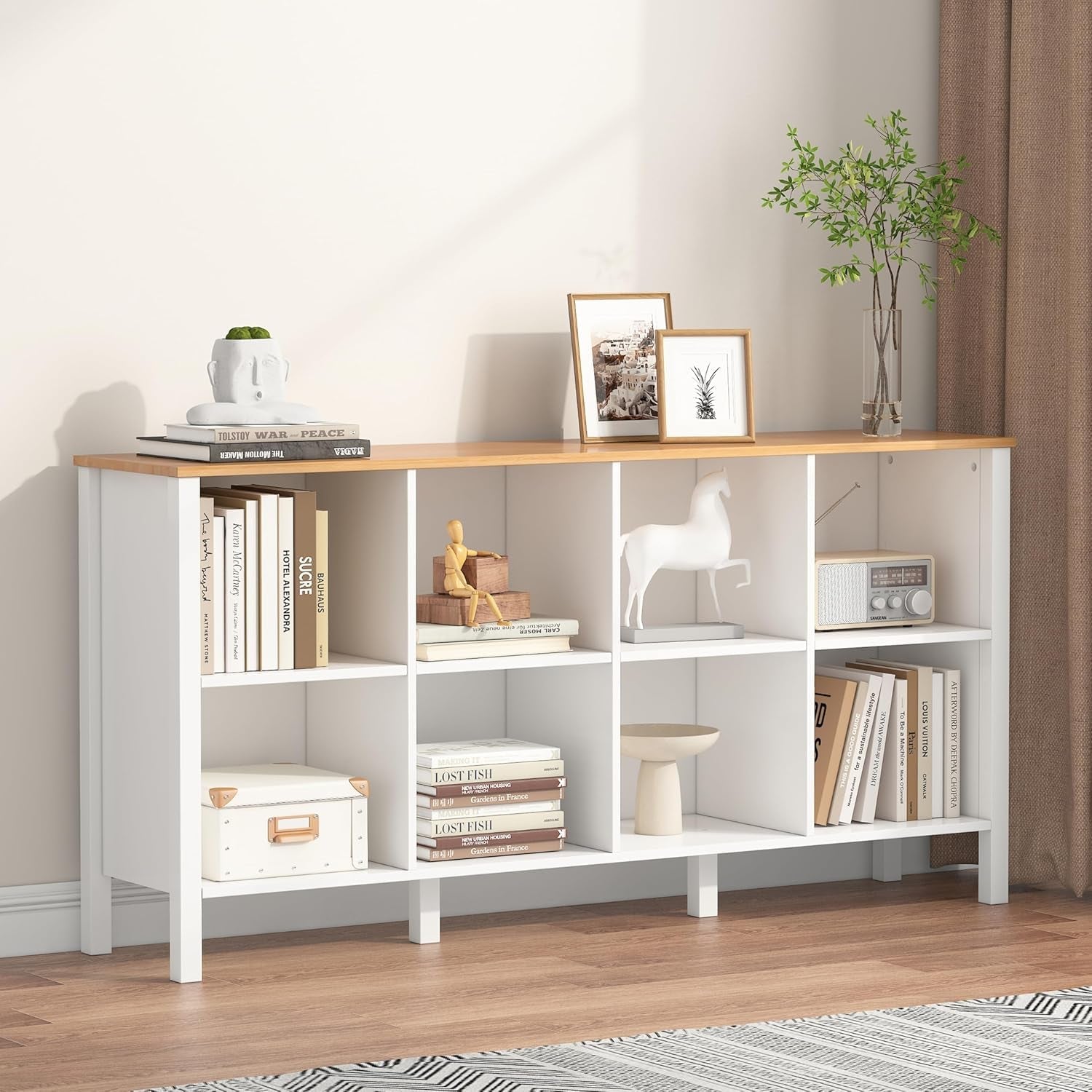 White bookshelf with wooden top against a neutral wall, displaying books, decor items, and plants.