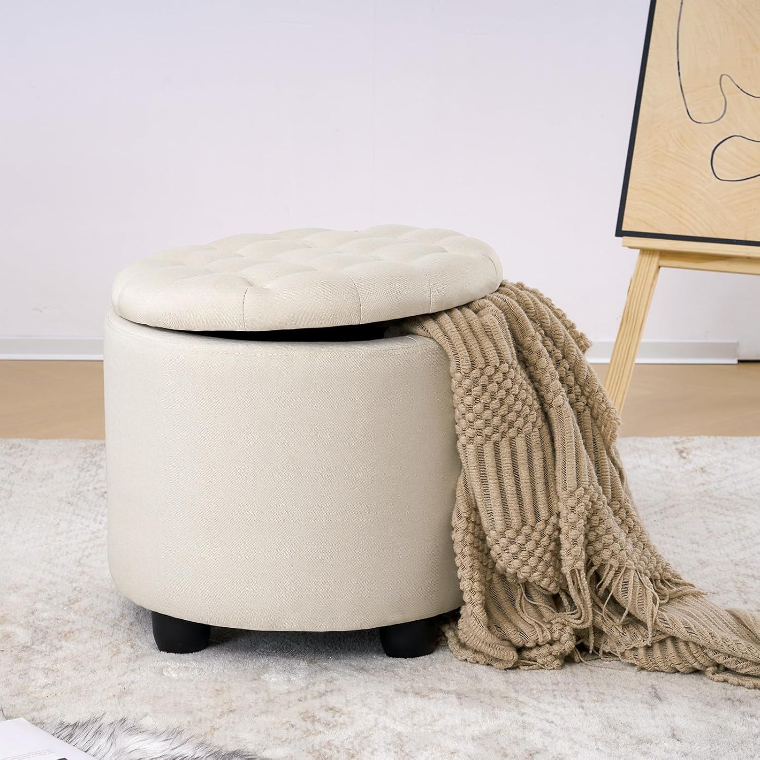 Beige ottoman with a blanket draped over it on a carpeted floor.