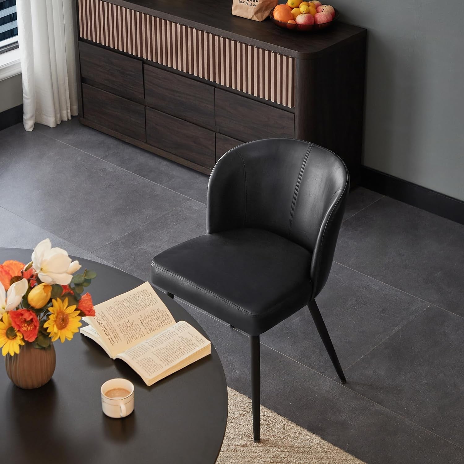 Black chair next to a round table with flowers, book, and cup in a room with dark wood furniture.