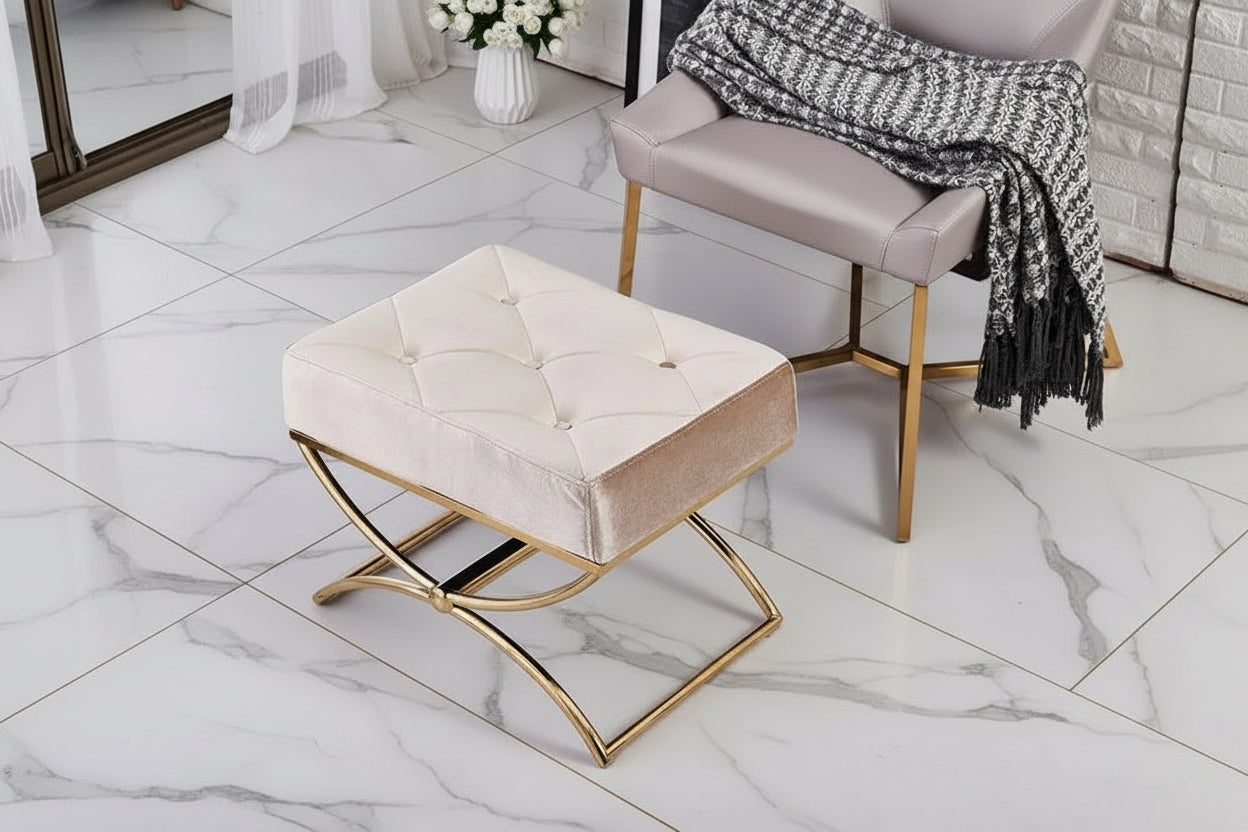Beige ottoman with gold legs on a tiled floor next to a chair with a blanket.