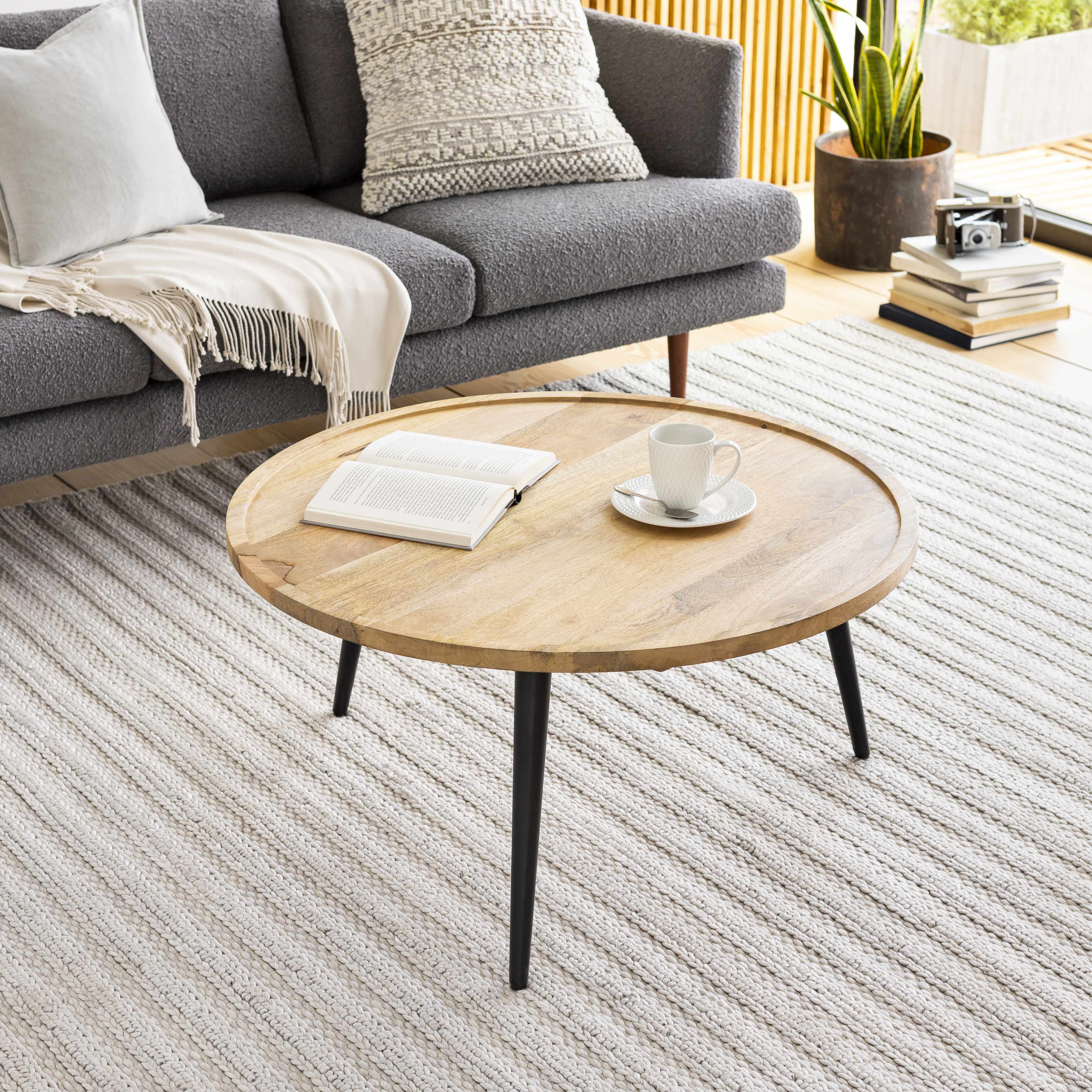 Round wooden coffee table in a living room with a gray sofa and books on a striped rug.