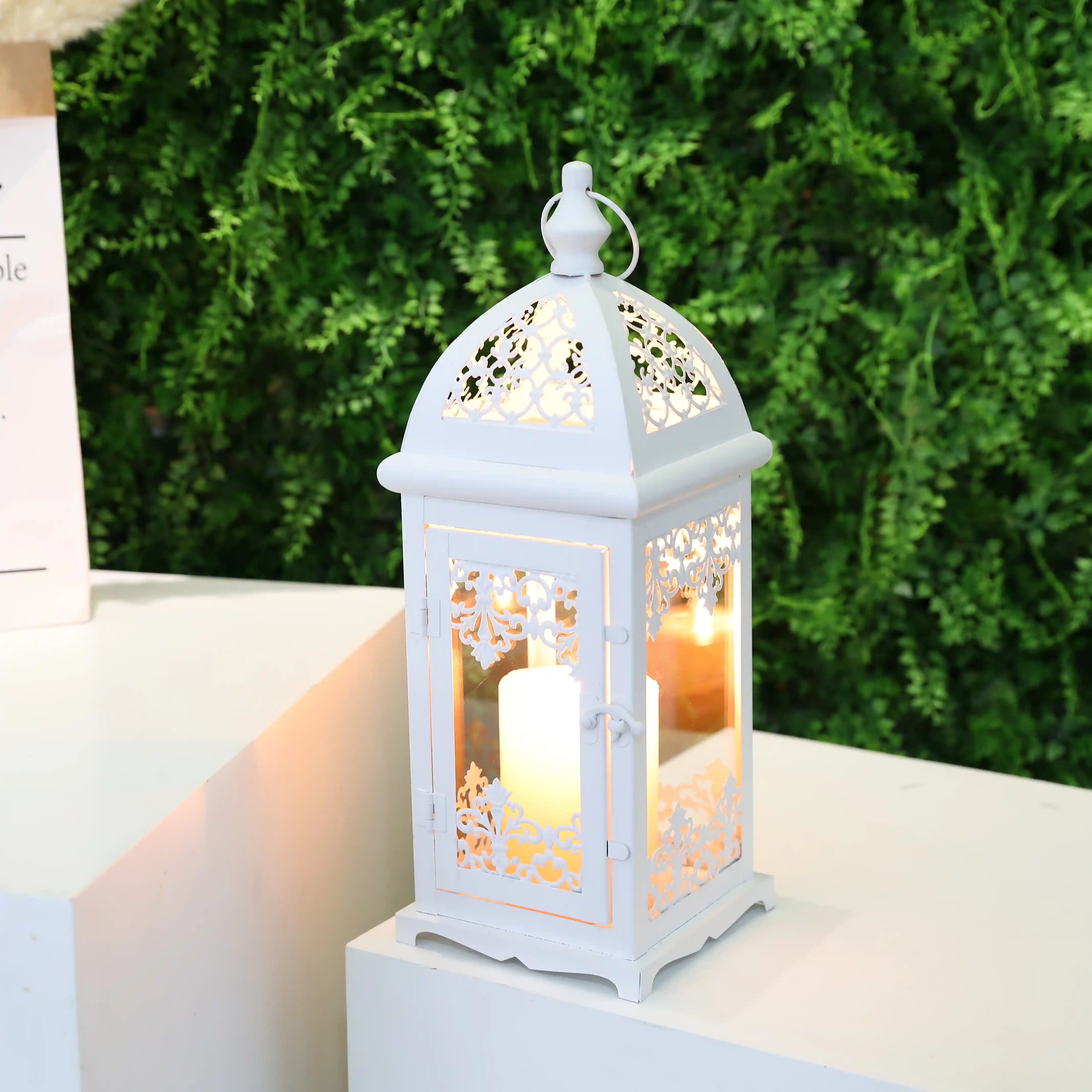 White decorative lantern with a lit candle inside on a white surface with greenery in the background