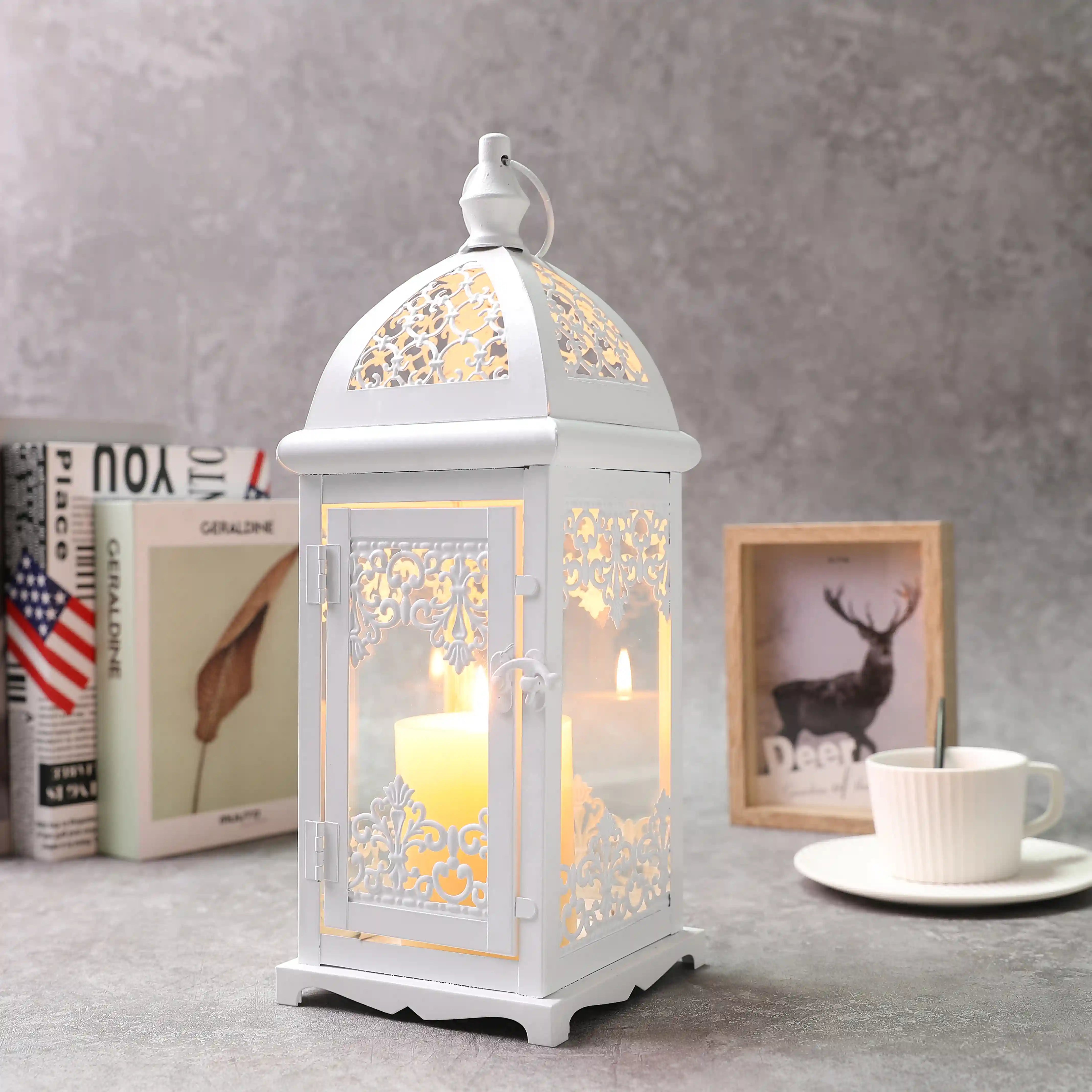 White decorative lantern with a lit candle inside on a table with books and a coffee cup in the background.