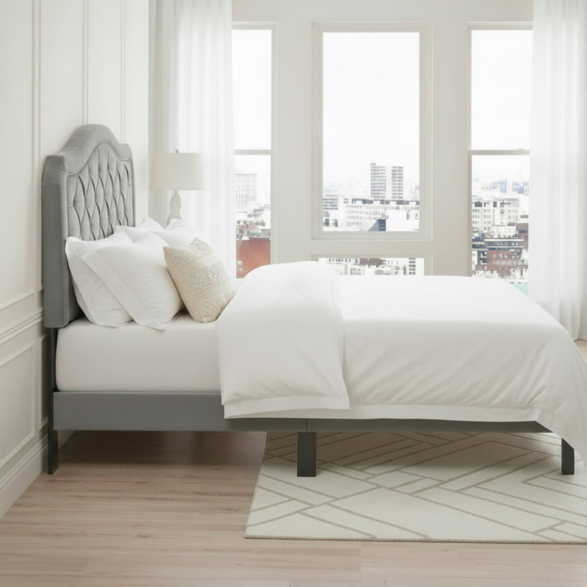 Gray upholstered bed with bedding in a room with large windows.