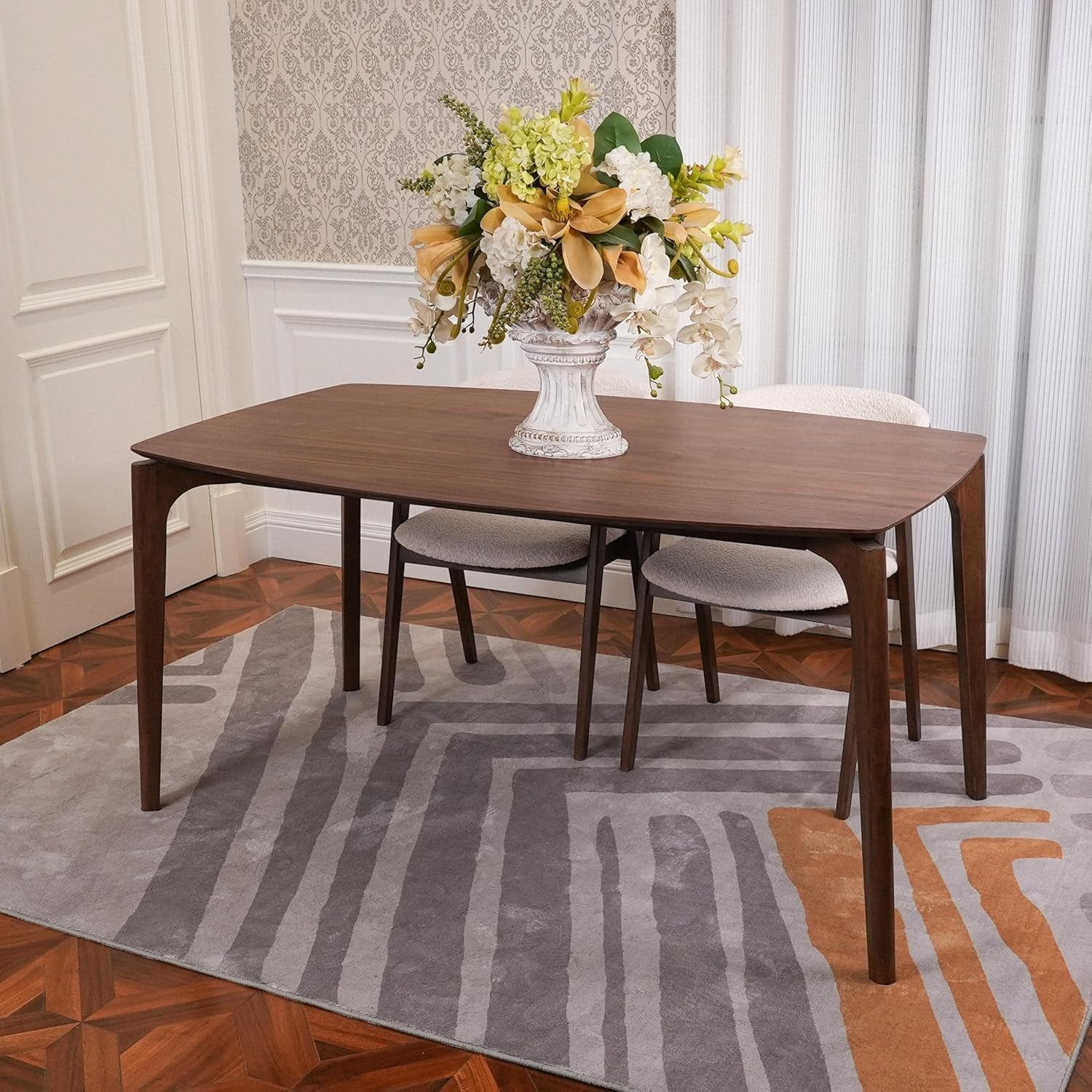 Wooden dining table with chairs and a floral arrangement in a room with patterned rug and wallpaper.