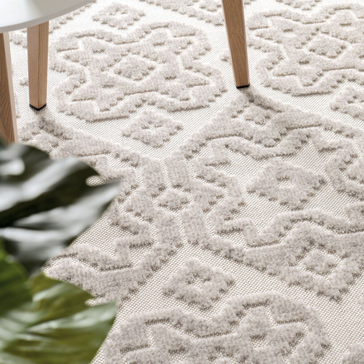 Close-up of a textured white rug with geometric pattern, partially covered by a plant.