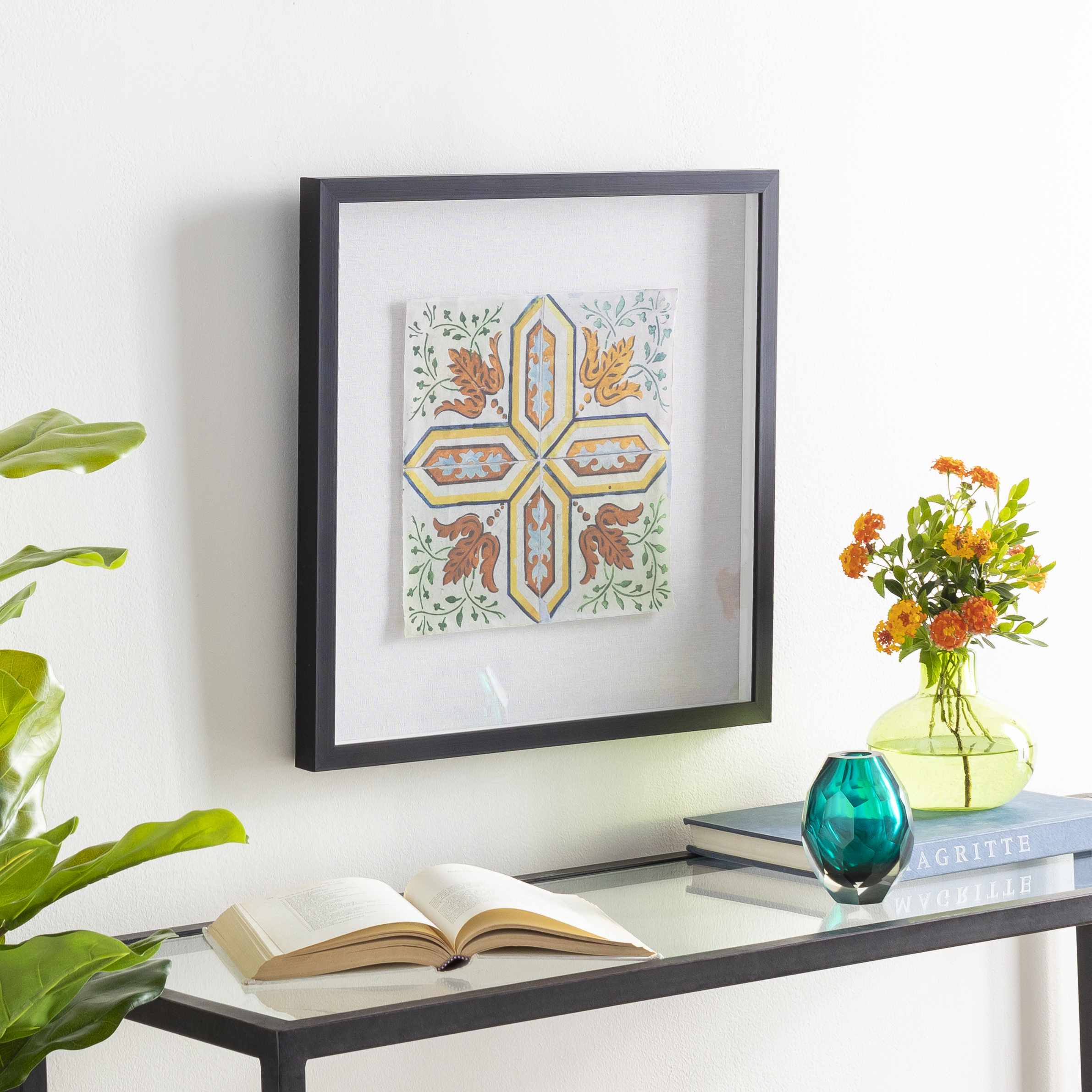 Framed artwork on a wall above a table with books and a vase.