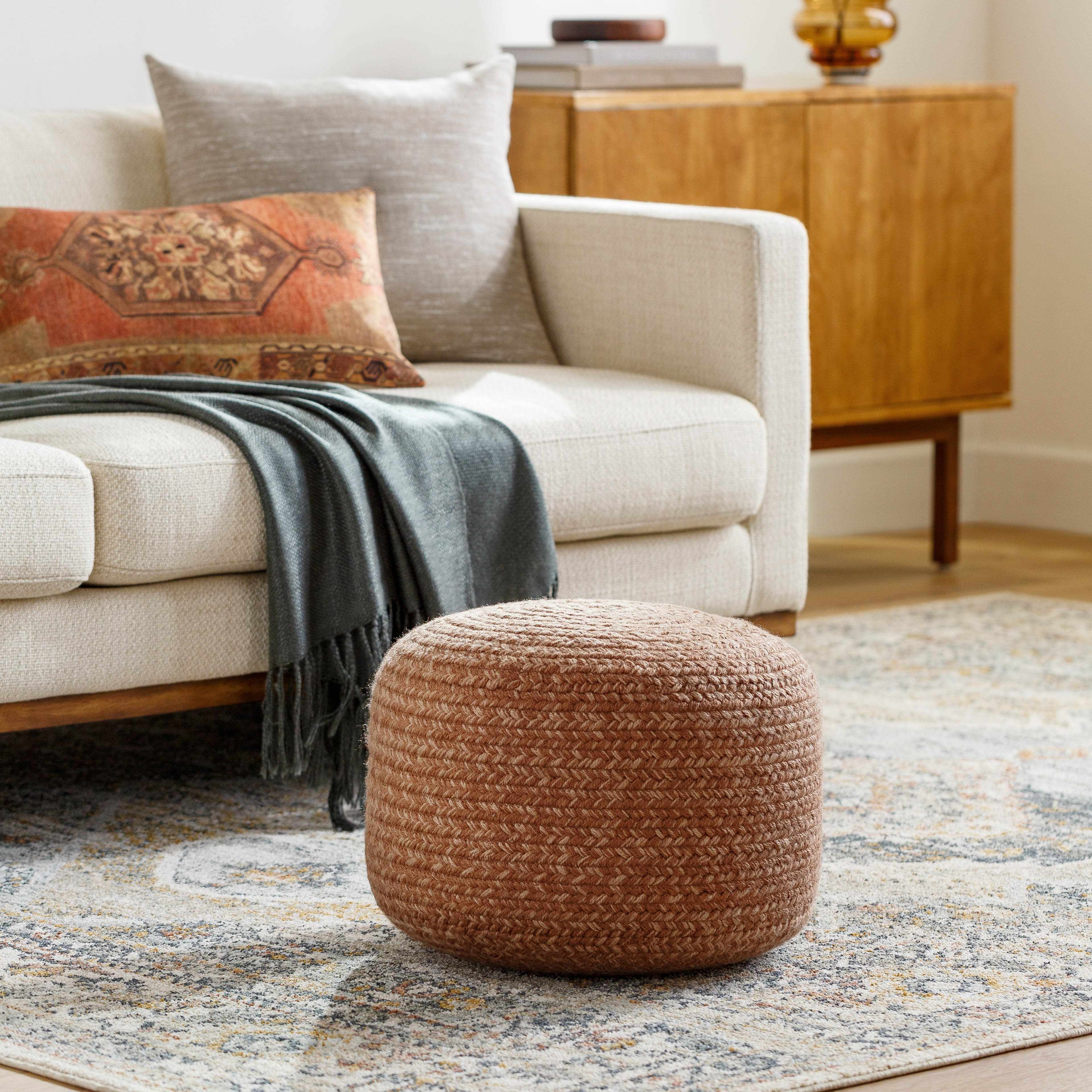 Brown woven ottoman in front of a beige sofa with decorative pillows and a wooden sideboard.