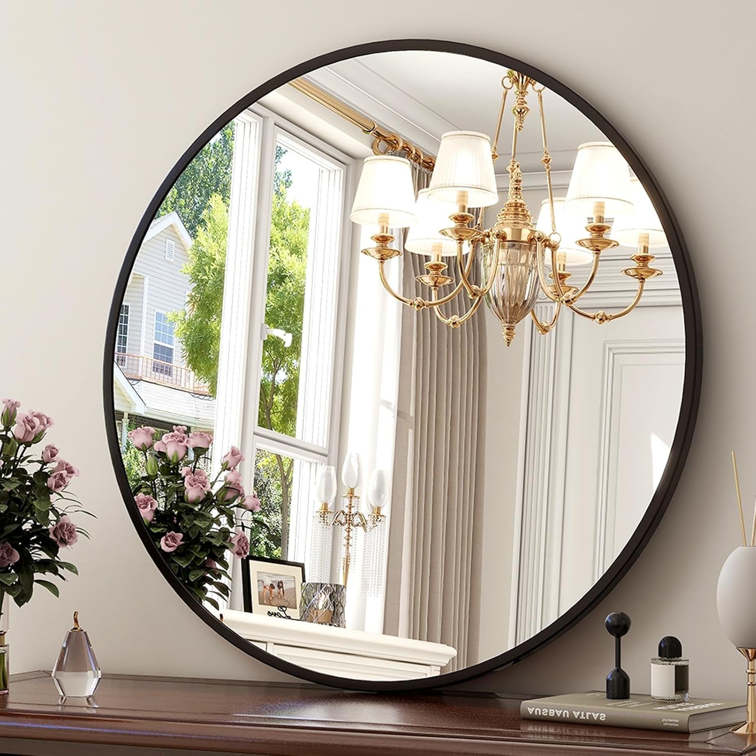 Round mirror on a dresser reflecting a chandelier and window with flowers.
