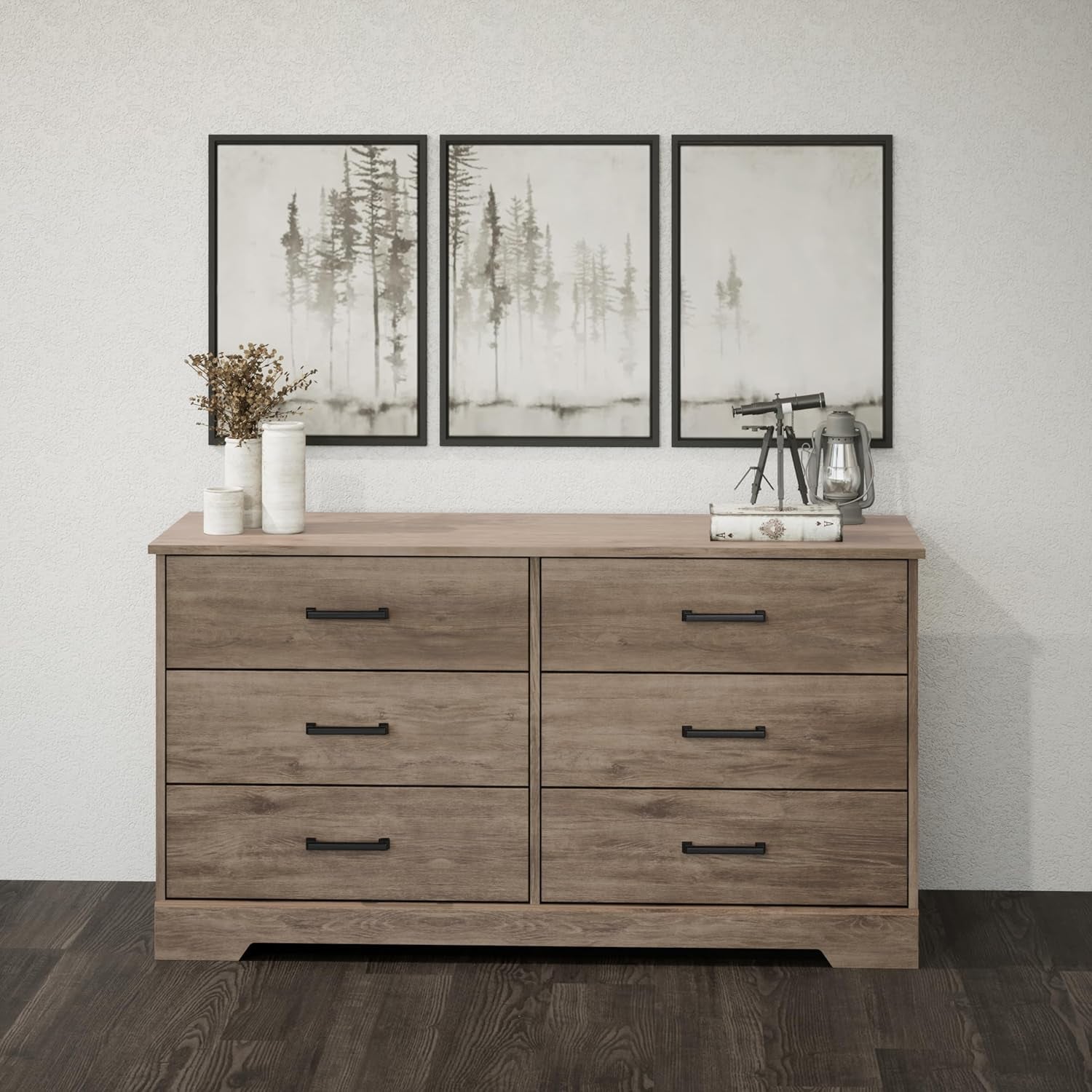 Wooden dresser with four drawers against a wall with three framed pictures.