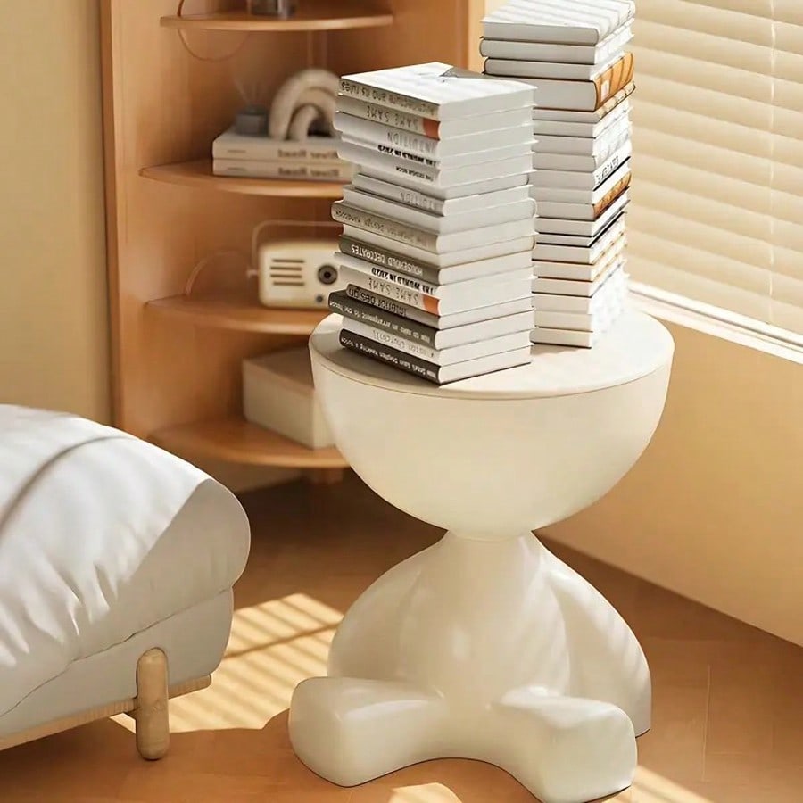 White side table with books on top next to a white chair in a room with wooden shelves.