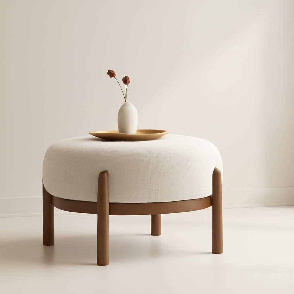 Beige ottoman with wooden legs in a room with a lamp and vase on a table.