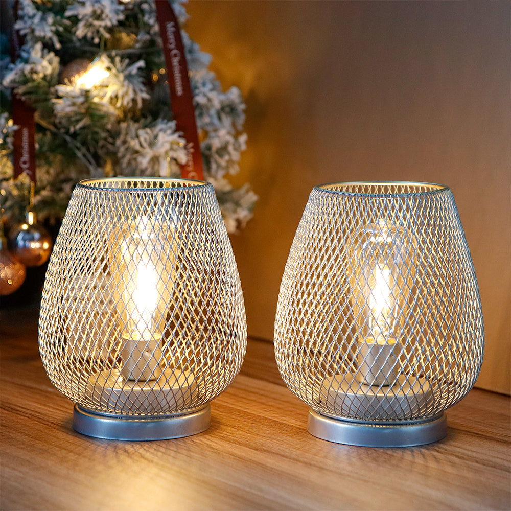 Two mesh-patterned candle holders on a wooden surface with a Christmas tree in the background.