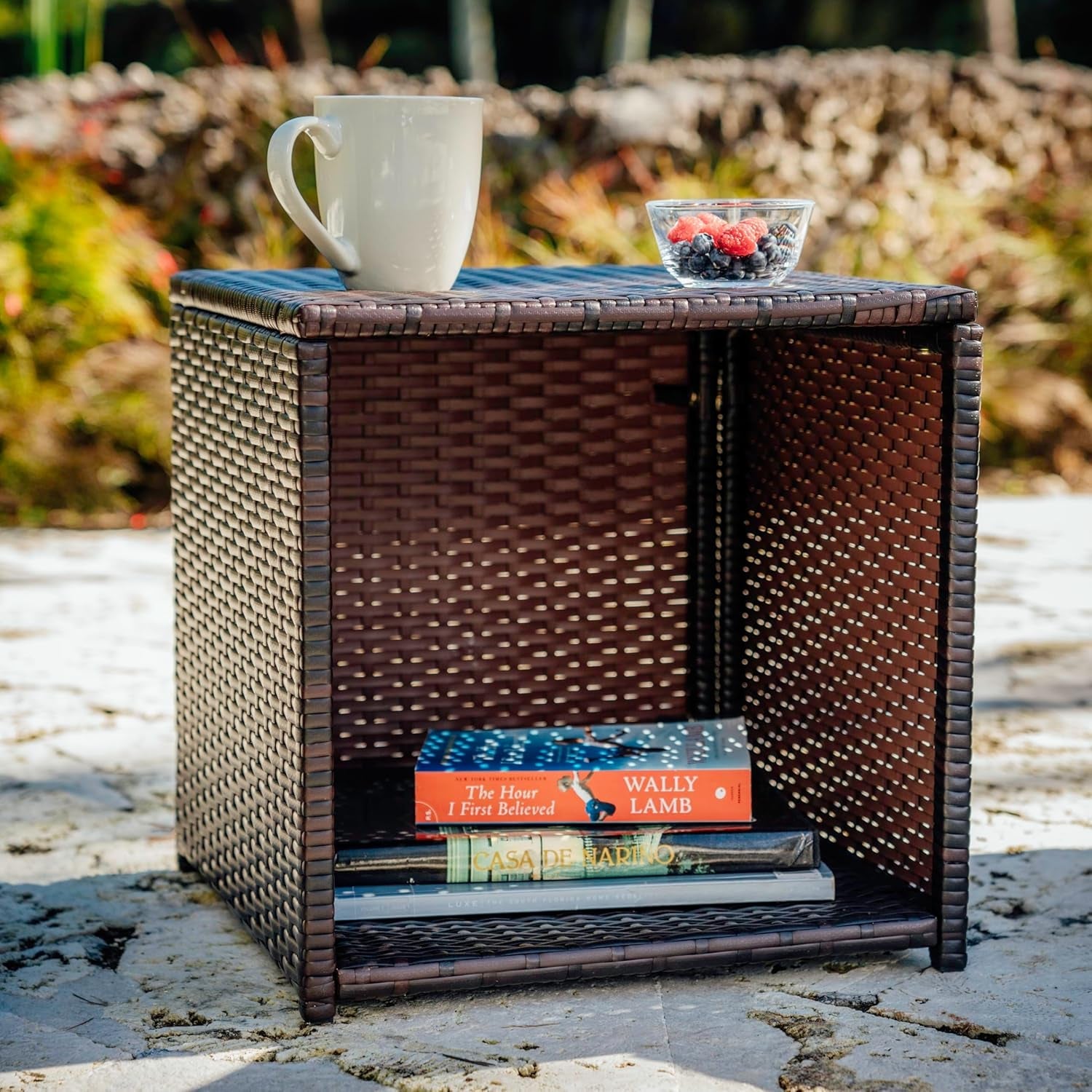 Wicker side table with a cup, bowl, and books on a stone surface outdoors.