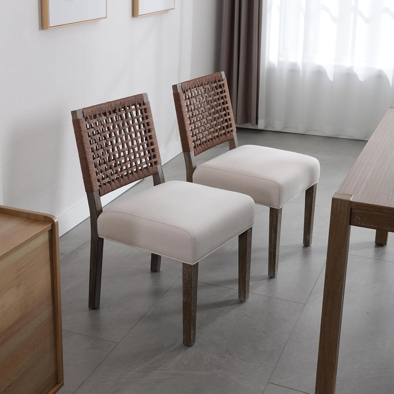 Two wooden chairs with beige cushions in a room with a table and curtains.