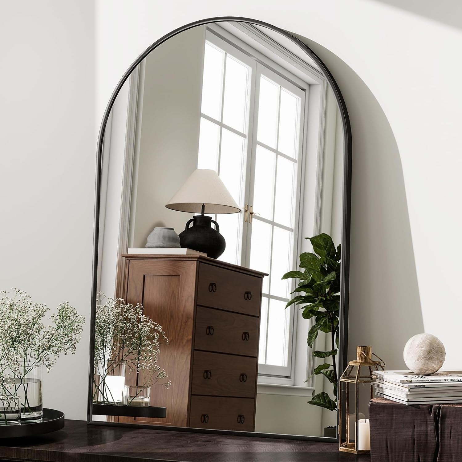 Large arched mirror reflecting a room with a wooden dresser, lamp, and plants.