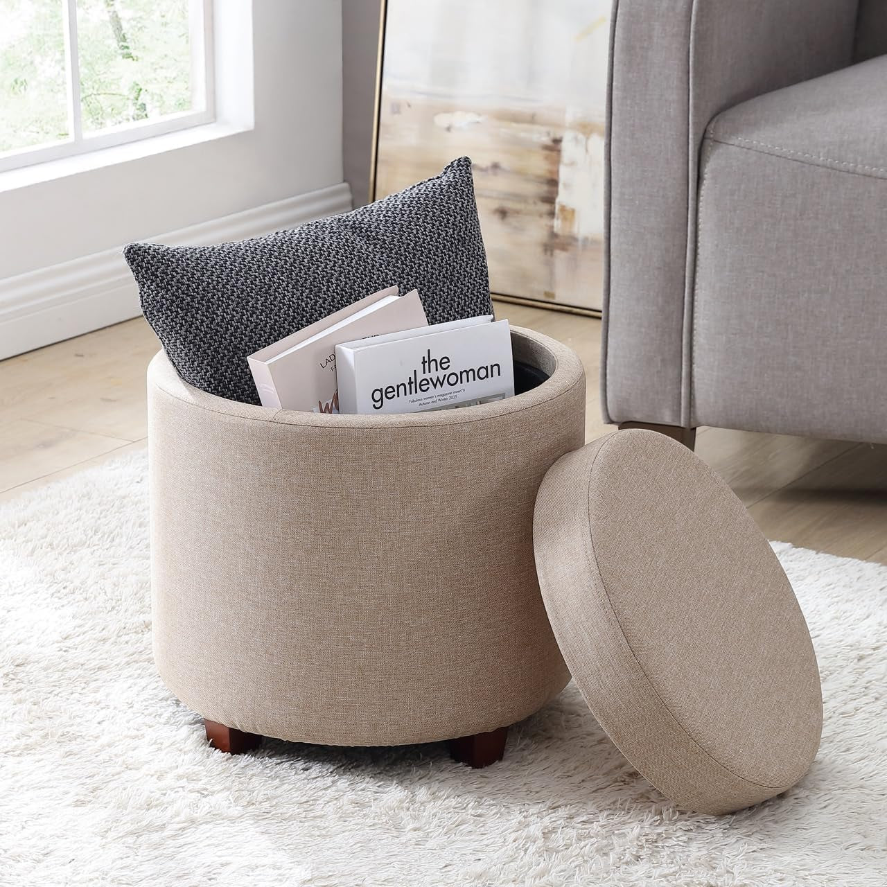 Round beige ottoman with a gray cushion and magazines on a white rug in a room.