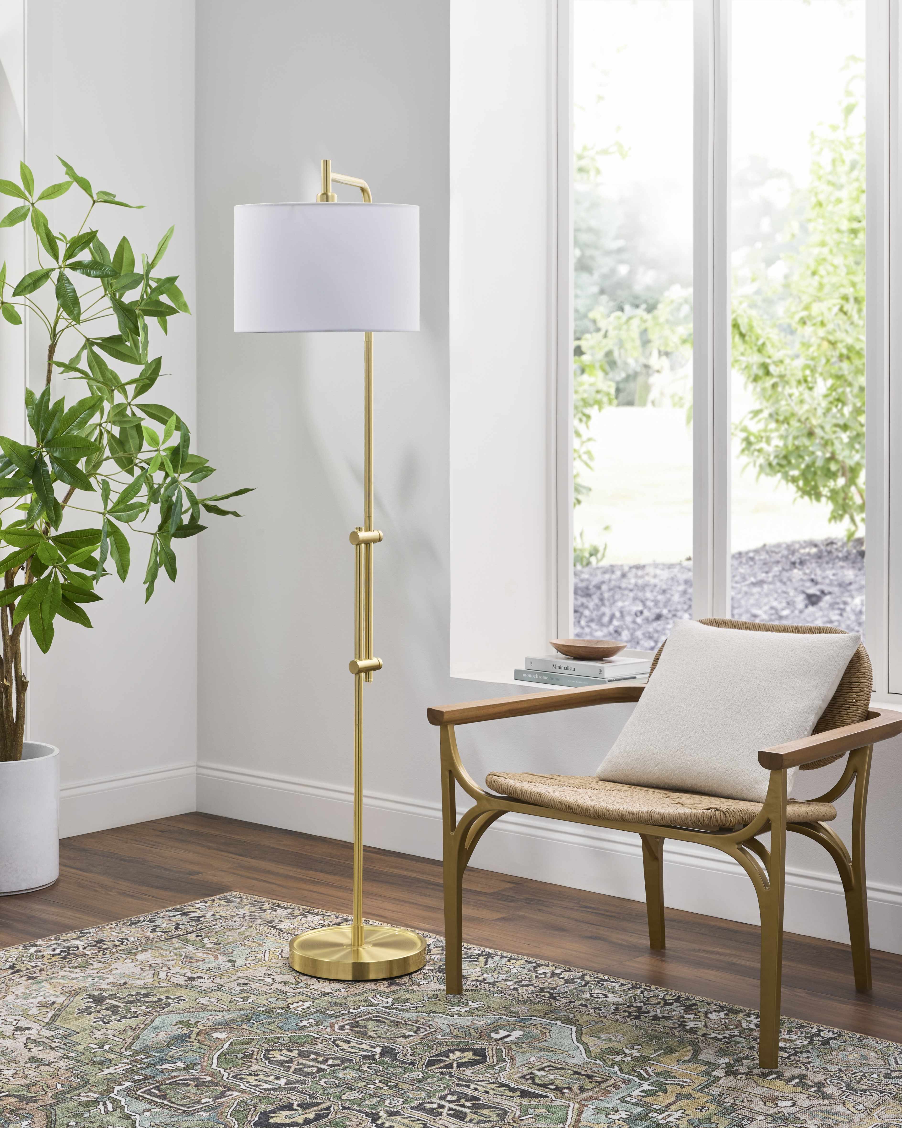 Gold floor lamp with white shade in a room with a chair, plant, and large window.