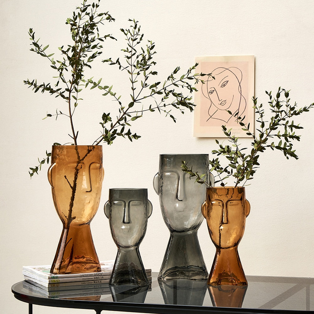 Set of four face-shaped glass vases with plants on a reflective surface.