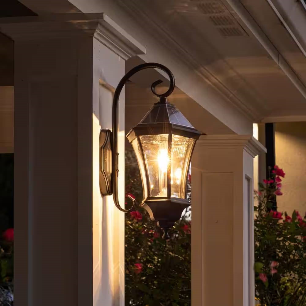 Decorative outdoor wall light fixture on a house exterior with flowers in the background.