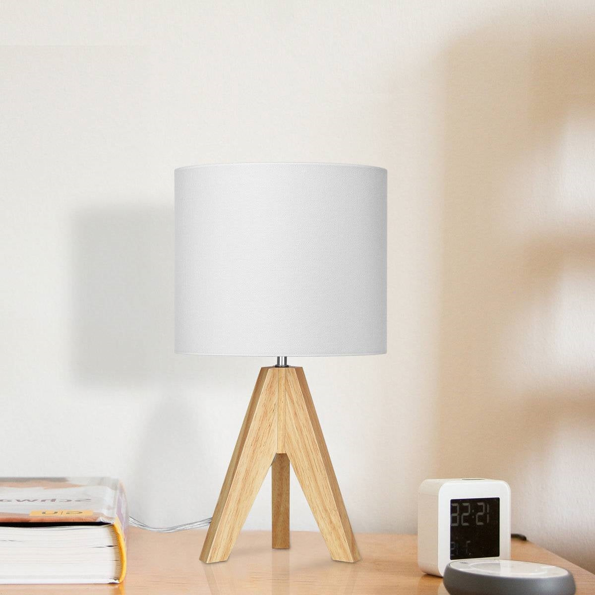Table lamp with wooden base and white shade on a desk next to books and a digital clock.
