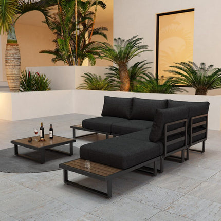 Outdoor patio set with black sectional sofa and wooden coffee table on a patio with plants in the background.