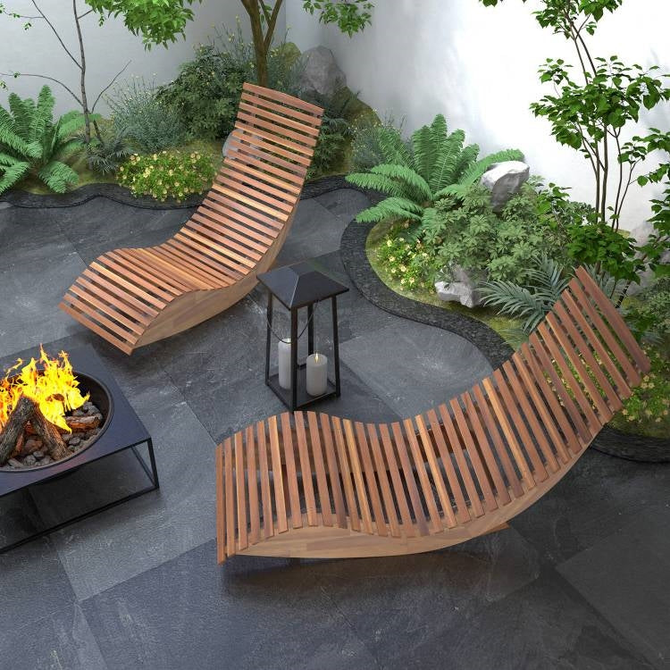 Two wooden lounge chairs and a fire pit in a garden setting with plants and rocks.