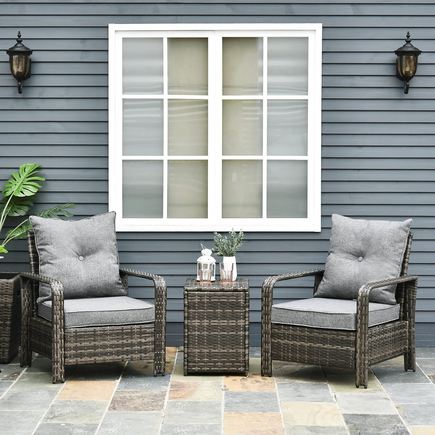 Two wicker chairs with gray cushions and a small table on a patio with a wall of color swatches in the background.