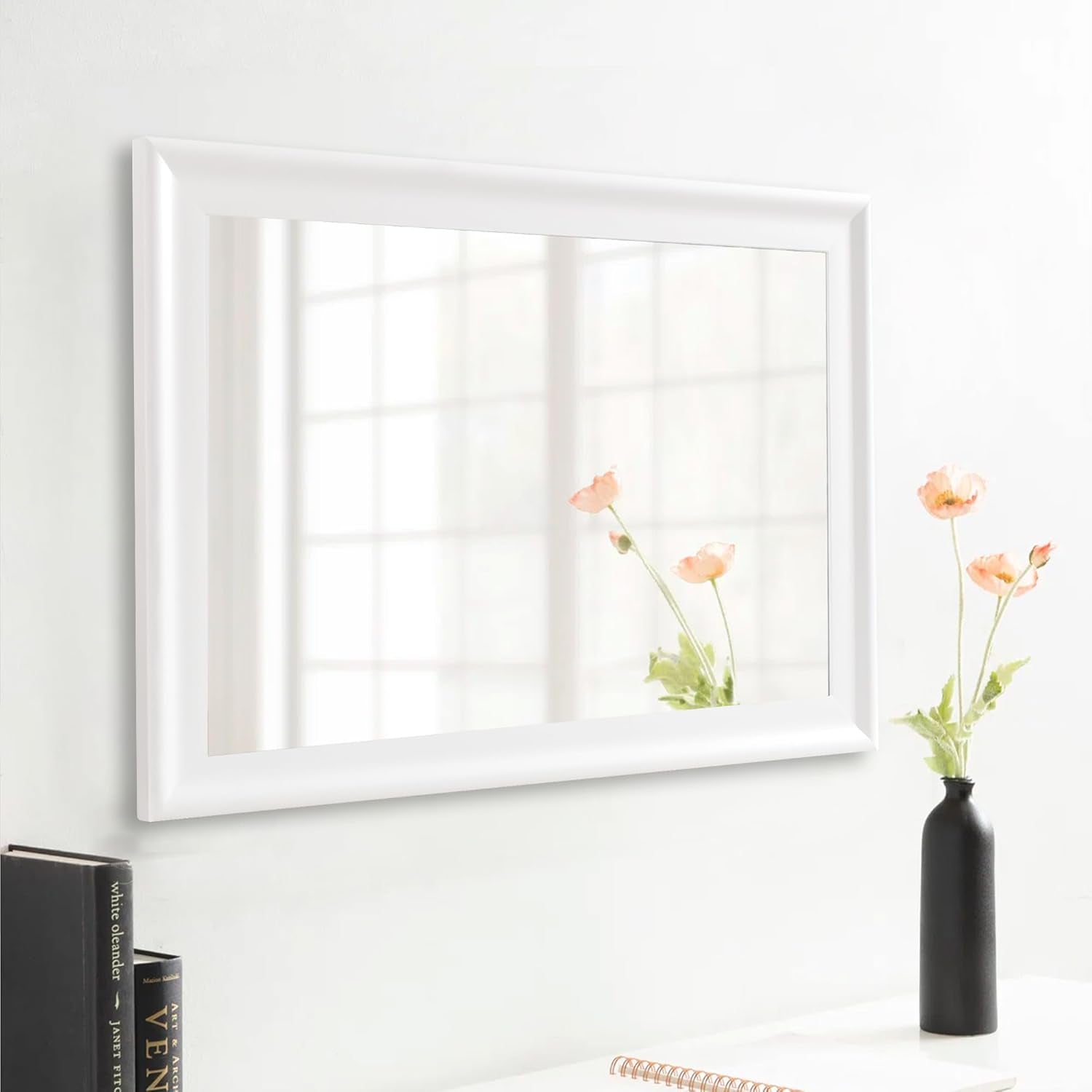 White framed mirror on a wall with a vase of flowers next to it.