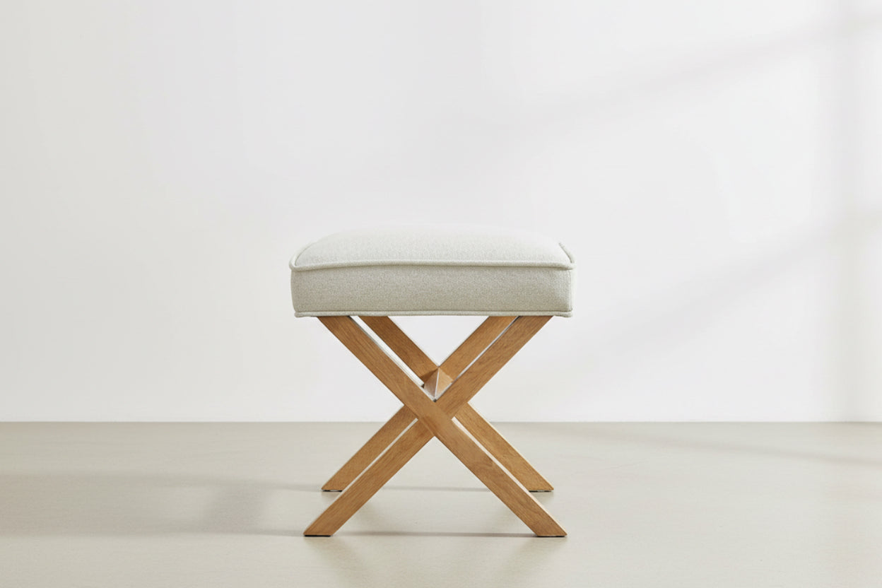 Stool with beige cushioned top and wooden cross-brace legs on a white background