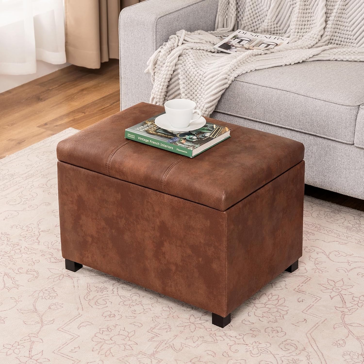 Brown leather ottoman with a cup and books on top, next to a gray sofa.