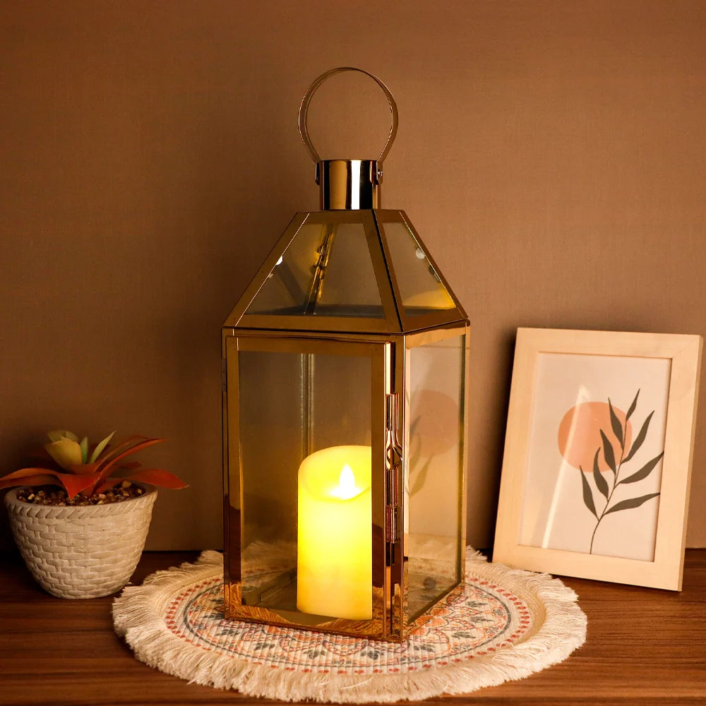Decorative lantern with a lit candle on a wooden surface, next to a potted plant and framed picture.