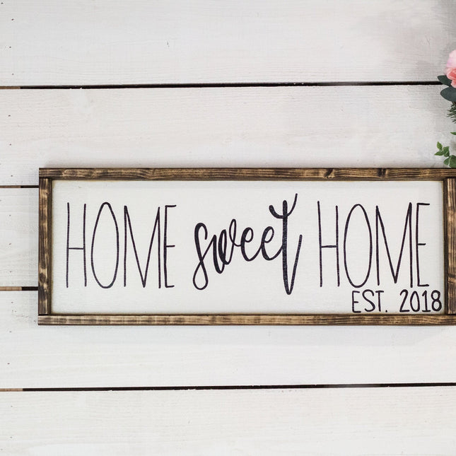 Decorative sign with 'Home sweet home est. 2018' on a white wooden background with flowers.